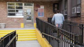 A Black, older resident uses a walker to get to the door of her public housing building, Wagner Houses