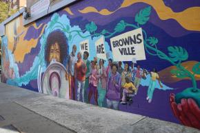 Partial view of mural featuring young woman speaking into a megaphone next to a group of people marching with garden tools and signs that read "We Are Brownsville."
