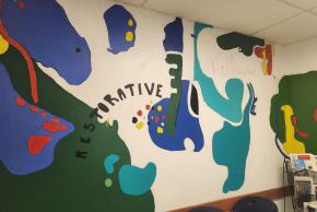 Indoor mural featuring various shapes of blue, red, green, and teal with the words "Restorative" and "Wellness" in decorative font on wall.