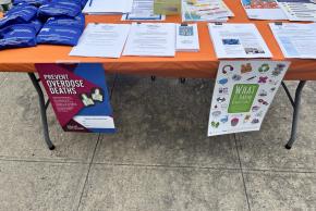 Outdoor table with overdose prevention kits, fentanyl testing strips, informational packets, and flyers reading "Prevent Overdose Deaths" and "What Is Harm Reduction?"