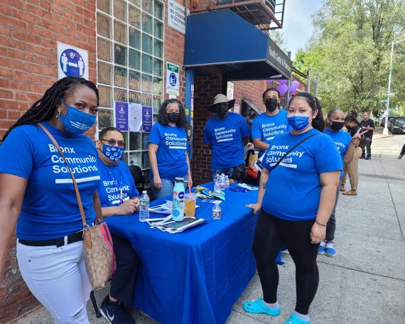 Bronx Community Solutions staff outside at an event; all dressed in Blue Bronx Community Solutions shirts and standing around a table with a blue tablecloth.