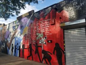 Memorial mural featuring names of deceased community members against a red backdrop with silhouettes of people.