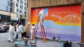 Group of painters working on colorful mural on outdoor wall featuring illustration of whale and text reading "Hope" and "We Love Far Rockaway."